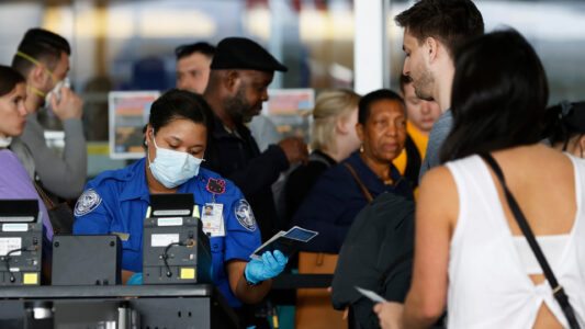 FILE - In this Saturday, March 14, 2020 file photo, a Transportation Security Administration agent hands a passport back to a traveler as she screens travelers, at a checkpoint inside an airline terminal at John F. Kennedy Airport in New York. The coronavirus pandemic that's caused many Americans to avoid airports has others booking spur-of-the moment trips at dirt-cheap ticket prices.  (AP Photo/Kathy Willens, File)